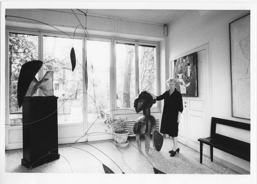 Peggy Guggenheim in the entrance hall of Palazzo Venier dei Leoni near a Yoka Mask (Nimba) from her collection of African artifacts, Venice, 1960s. Solomon R. Guggenheim Foundation. Photo Archivio CameraphotoEpoche. Gift, Cassa di Risparmio di Venezia, 2005 Left to right, Antoine Pevsner, Developable Surface (1941, PGC), Alexander Calder, Arc of Petals (1941, PGC), Georges Braque, The Bowl of Grapes (1926, PGC).